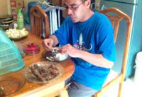 Di meja makan di dapur, saya mencicipi ayam panggang buatan istri saya. Istri saya memotret sambil juga menggerogoti paha ayam kesukaannya