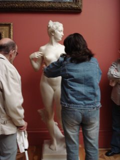 A woman touching the side of a sculpture of a young nude woman, and Ken waiting his turn