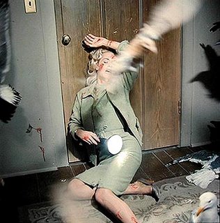 Tippi Hedren está caída no chão, com o corpo encostado em uma porta fechada, com marcas de arranhados e sangue espalhados por rosto, mãos, braços e tornozelo, e tenta se defender de pássaros que a atacam.