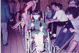 Me, aged 5, on a swimming pool side in my first, red, wheelchair, with my left leg in a full length cast, clutching a trophy